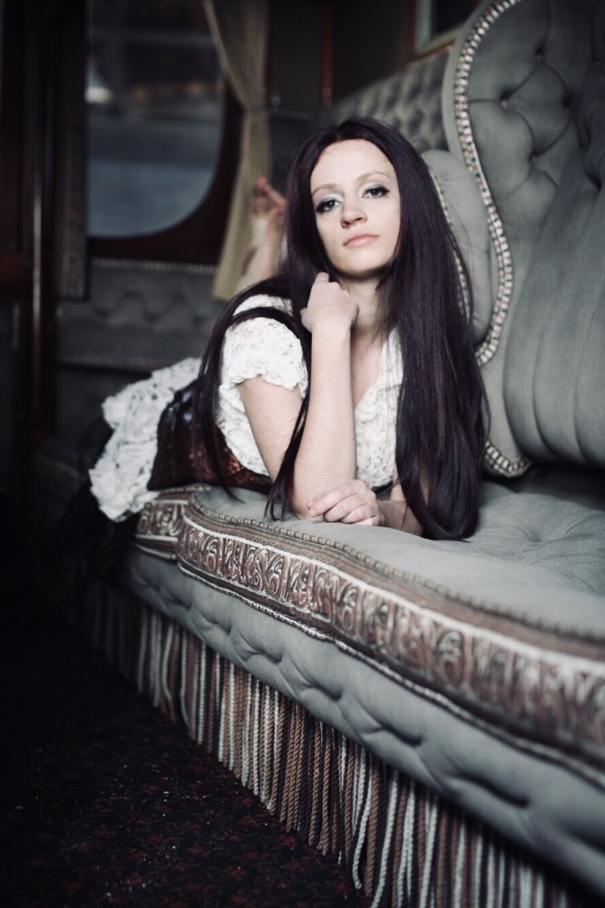 femme gothique en costume steampunk et cheveux longs allongée sur le ventre portrait sur une banquette grise ancienne
