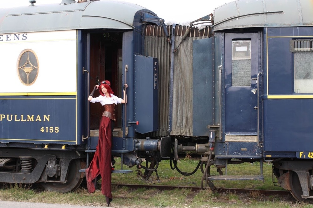 femme gothique en costume steampunk et cheveux longs devant la porte d'un wagon bleu ancien