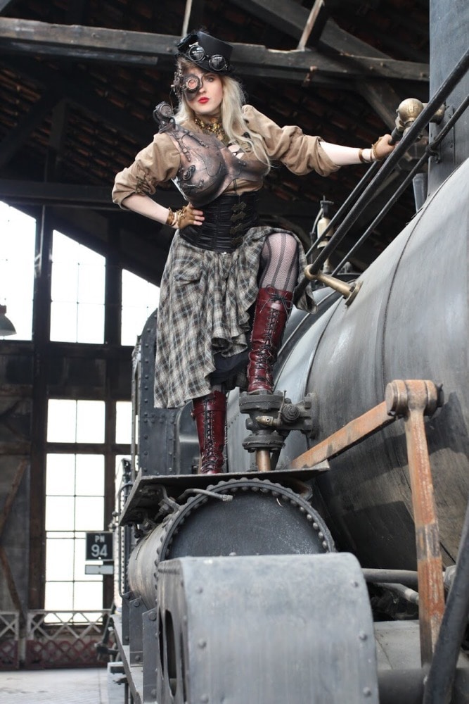 femme en costume steampunk à côté d'une ancienne locomotive dans un hangar