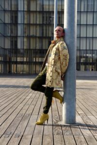 Homme en long manteau jaune et chaussures jaunes debout contre un poteau sur le parvis de la BNF Paris