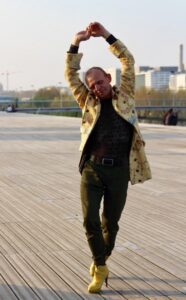 Homme en manteau long jaune et talons hauts jaunes dansant sur le parvis de la BNF