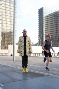 Homme en long manteau jaune et chaussures à talons jaunes devant les tours de la grande bibliothèque Paris BNF