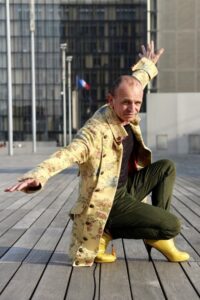 Homme en manteau long jaune et talons hauts jaunes accroupis en faisant l'avion sur le parvis de la BNF