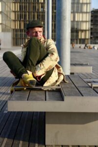 Homme en manteau long jaune et talons jaunes allongé sur un banc public