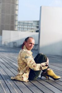 Homme en manteau long jaune et talons jaunes assis sur le parvis de la BNF Paris