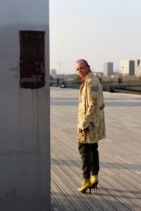 Homme en manteau long jaune et talons hauts jaunes debout devant un mur du parvis de la BNF