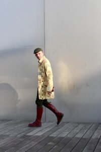 Homme lifestyle au manteau long jaune et bottes rouges marchant devant un mur brillant