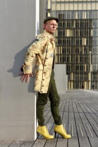 Homme en manteau long jaune et talons hauts jaunes contre un mur de profil sur le parvis de la BNF