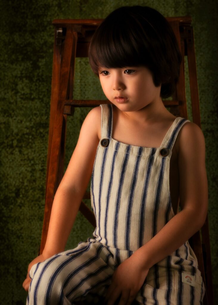 portrait enfant eurasien en clair obscur en salopette sur un escabeau en bois