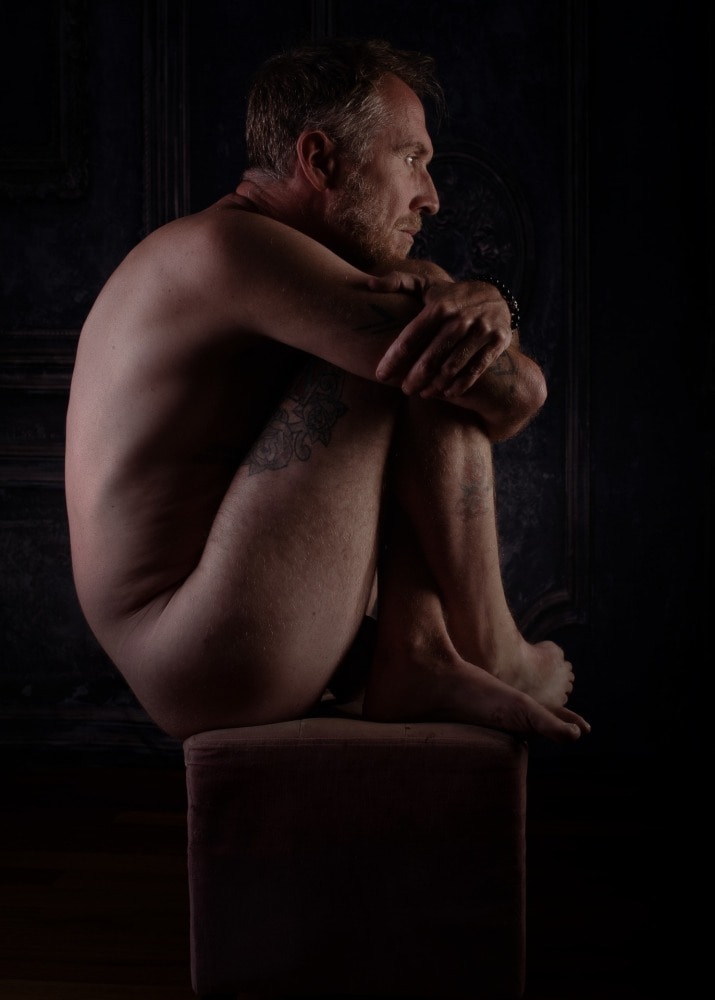 homme barbu et tatoué assis de profil en boule sur un tabouret
