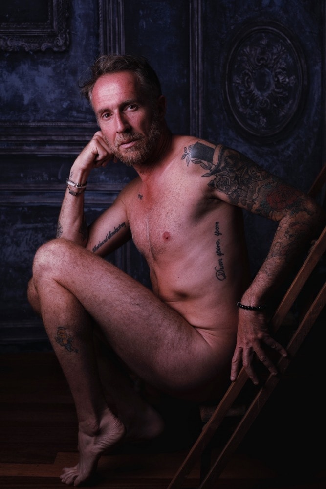 homme tatoué en nu artistique clair obscur de profil assis sur un escabeau