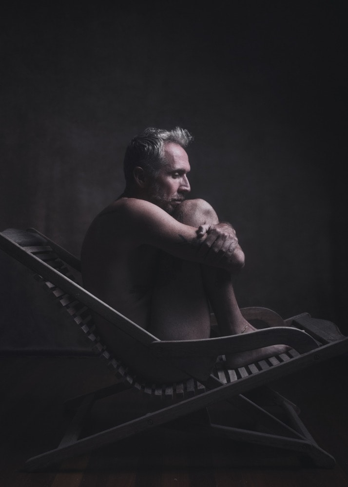 homme en nu artistique clair obscur assis sur une chaise longue en bois