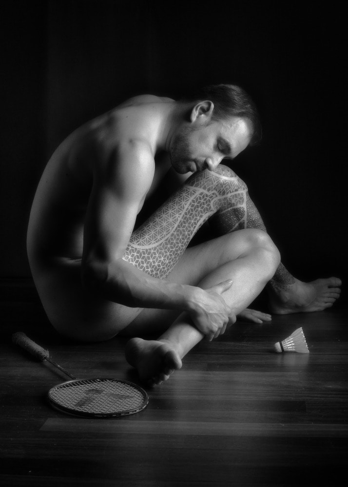 homme en nu artistique assis au sol recroquevillé sur une jambe tatouée atrophiée