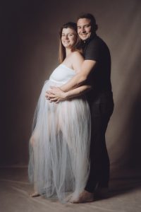 Séance couple enceinte estime avec Sarah et Benoît robe blanche tulle debout mains sourire sépia par femme photographe grossesse au studio photo location Céline Pivoine Eyes à Montreuil 93 près de Vincennes