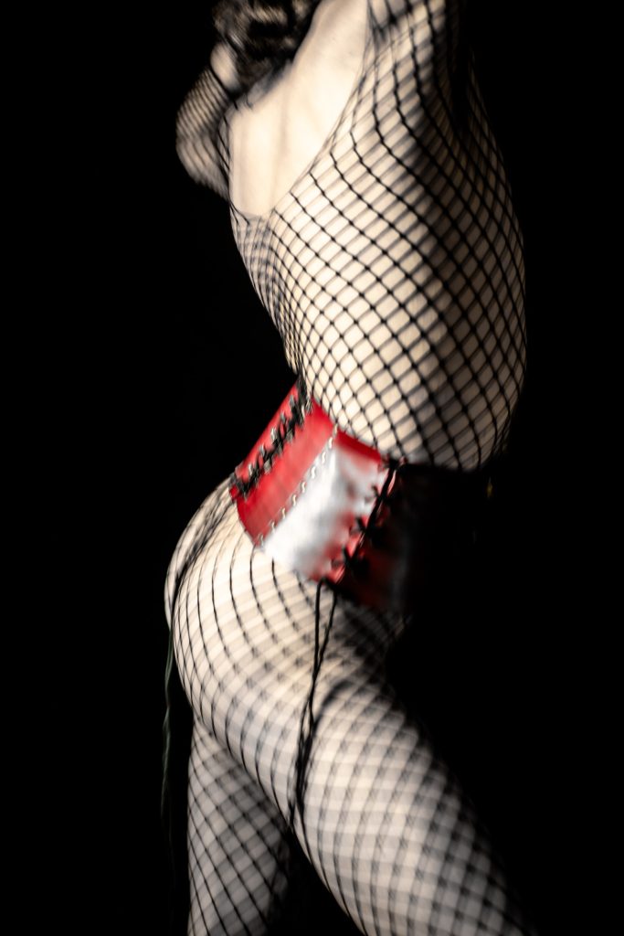 Séance photo shooting boudoir pour homme féminin avec Dione par femme photographe Céline Pivoine Eyes au studio photo location à Montreuil 93 près de Vincennes serre taille rouge cuir et résilles profil debout