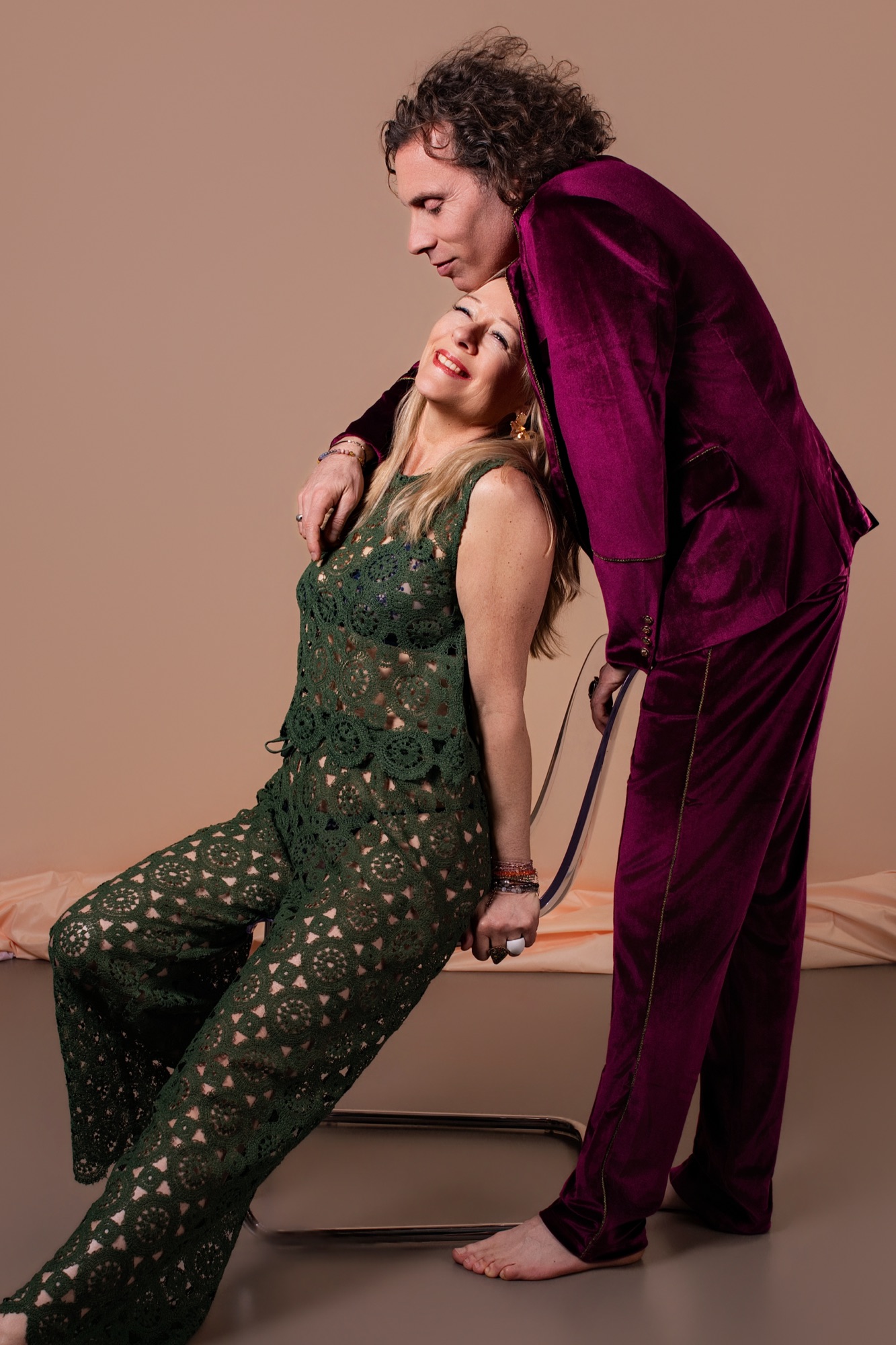 Portrait de couple élégant en studio — elle assise sur une chaise en combinaison crochet verte renversée en arrière souriant, lui penché au-dessus d'elle en costume velours bordeaux la main sur l'épaule — femme photographe Céline Pivoine Eyes au studio photo à Montreuil près de Noisy-le-Sec