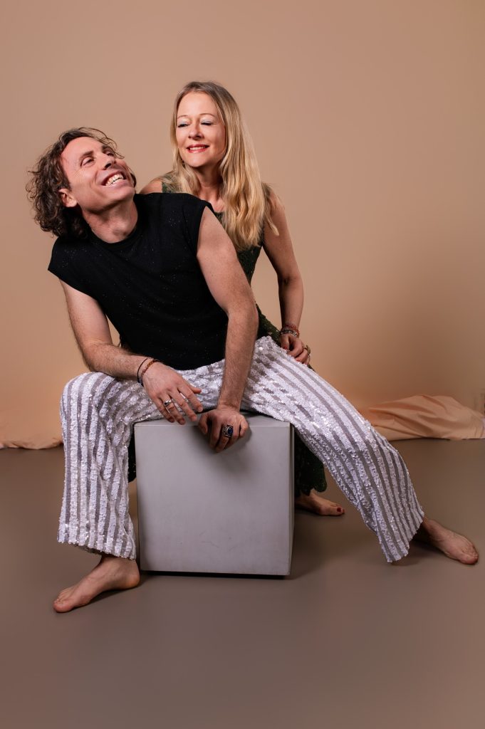Portrait de couple complice en studio — il rit aux éclats assis sur un cube, elle appuyée dans son dos souriante, tous deux pieds nus en pantalons à sequins — femme photographe Céline Pivoine Eyes au studio photo à Montreuil près de Noisy-le-Sec