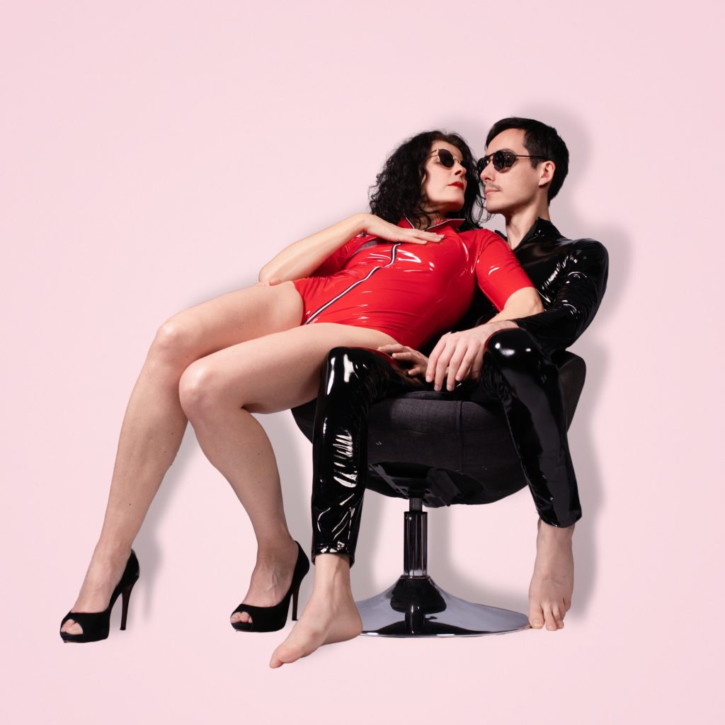 Couple enlacé sur un fauteuil pivotant noir, elle assise sur ses genoux dans une combinaison vinyle rouge à zip, jambes croisées en escarpins ouverts noirs, lui en tenue vinyle noire pieds nus, tous deux portant des lunettes de soleil rondes sur fond rose pastel, séance photo duo par la femme photographe Céline Pivoine Eyes au studio photo à Montreuil près de Noisy-le-Sec.