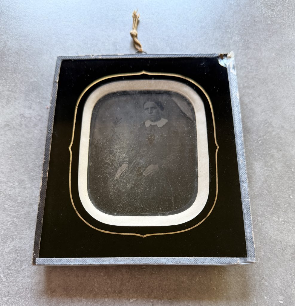 Ambrotype sous cadre noir laqué à filet doré : portrait d'une femme d'âge mûr au grand col blanc arrondi, devant un décor de feuillage peint, vers 1855-1865.