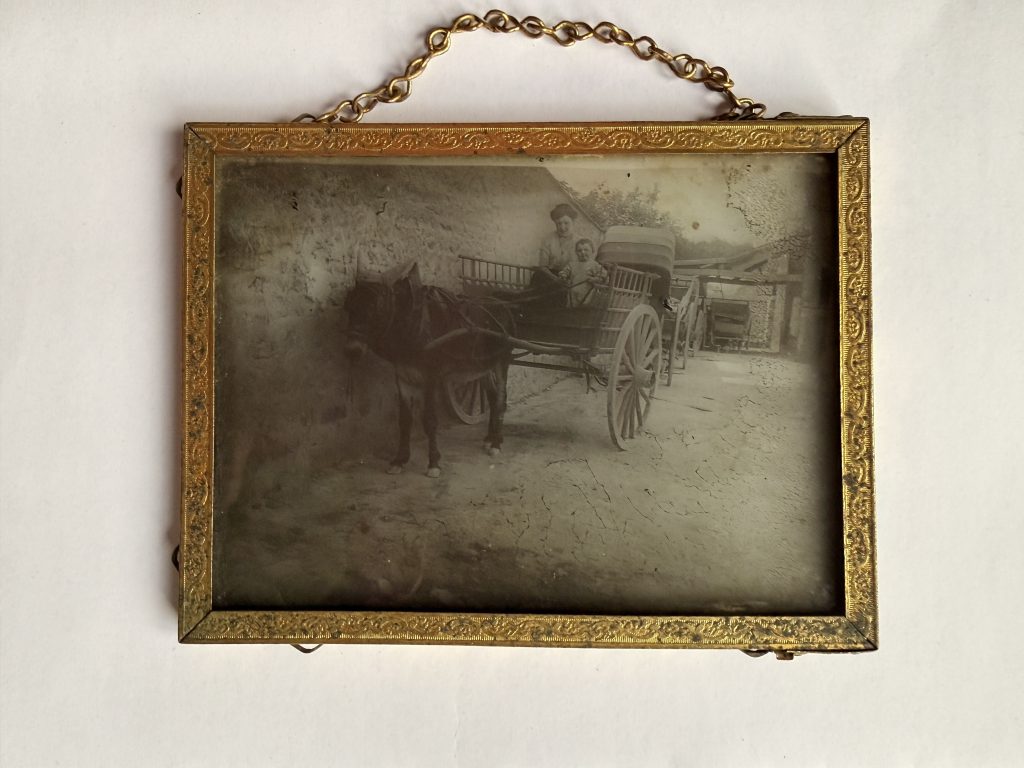 Positif photographique sur plaque de verre dans un cadre en laiton doré : une femme au grand chapeau et un jeune enfant en robe blanche dans une carriole attelée à un âne, scène de cour de ferme, vers 1905-1915.